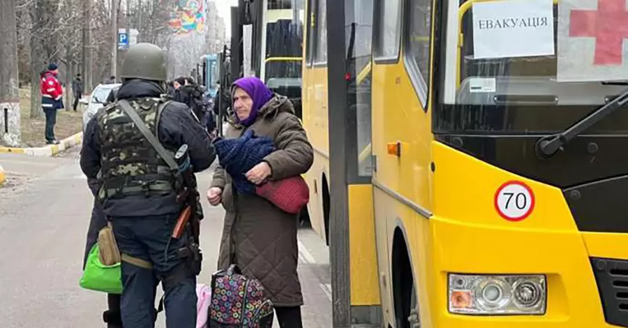 Військовий допомагає літній жінці з речами біля жовтого евакуаційного автобуса