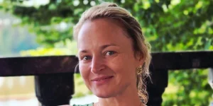 Жінка з світлим волоссям усміхається на тлі зеленого листя та деревяної огорожі