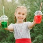 Улыбающаяся девочка с косами держит два цветных напитка в стаканах