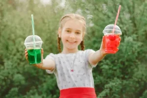 Усміхнена дівчинка з косами тримає два кольорові напої в стаканах