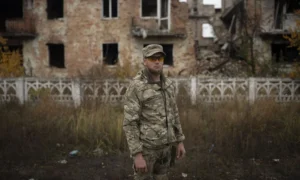 Чоловік у камуфляжі та жовтих окулярах стоїть перед зруйнованою будівлею